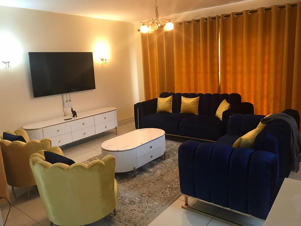 A spacious living area features a large flat-screen TV mounted on the wall. Navy blue sofas with yellow cushions are arranged around a coffee table. Yellow curtains provide a warm backdrop, complemented by elegant wall lighting and a patterned area rug on the tiled floor.