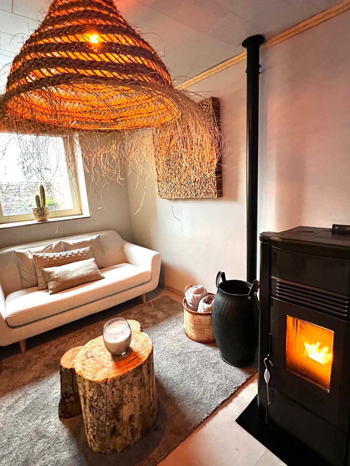 A cozy sitting area features a comfortable white sofa positioned beside a wood-burning stove. A soft rug lies beneath, complemented by a rustic wooden stump as a coffee table. Natural light filters in through a window adorned with plants, enhancing a peaceful ambiance.