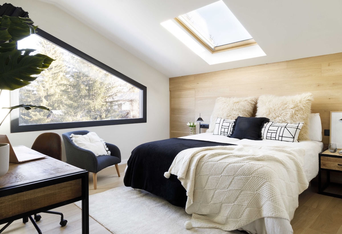 A bright sleeping area is defined by a spacious bed featuring neutral bedding with decorative pillows. Soft textures are introduced through a knitted throw. A large angled window provides natural light and views of greenery, while a seating area includes a cozy chair and small table.