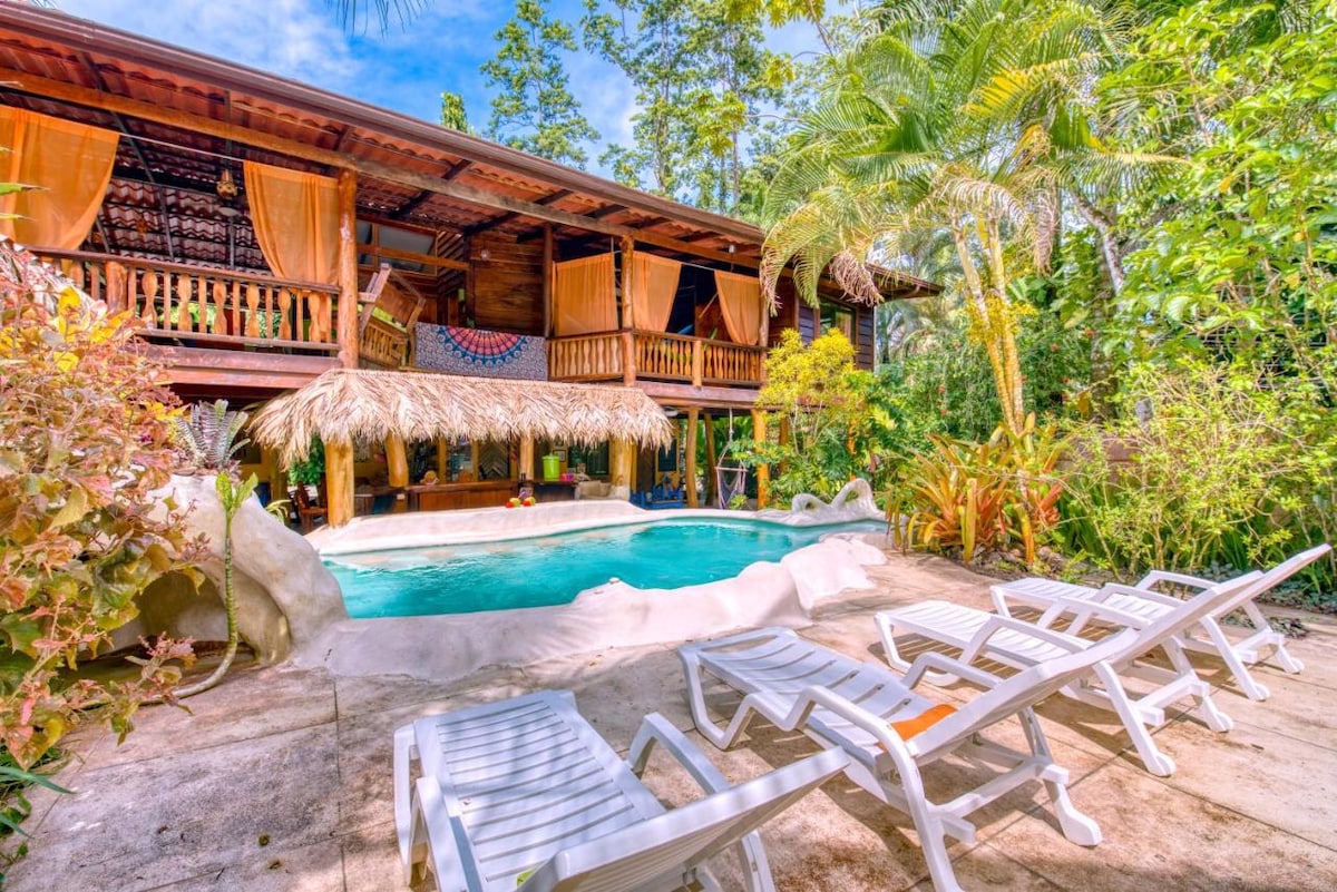 A tropical retreat features a swimming pool surrounded by lush greenery, complemented by a thatched-roof structure in the background. Multiple lounge chairs are positioned near the pool, providing spaces for relaxation. Colorful foliage enhances the natural atmosphere of the setting.