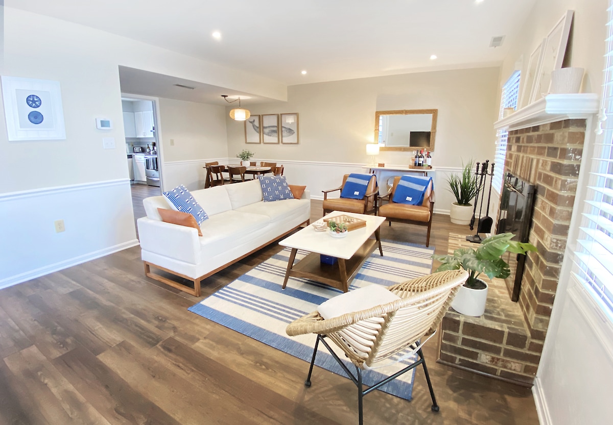 A modern living area features a white sofa with wooden accents, accompanied by two wooden armchairs. A light blue area rug lays on the hardwood floor, and a coffee table is set on top. Natural light filters through the windows, enhancing the space.
