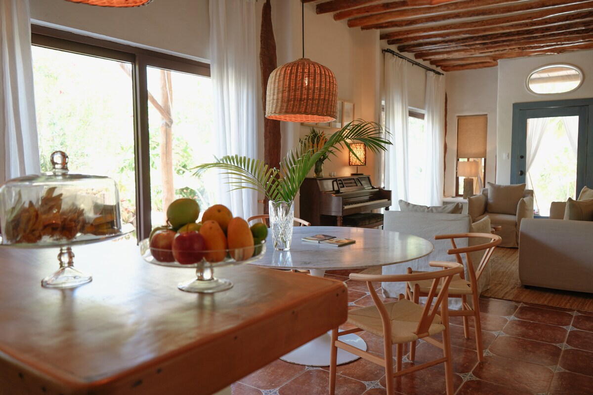 A dining area is presented with a circular wooden table surrounded by light-colored chairs. Fresh fruit is displayed in a glass bowl at the center. Natural light enters through large windows, complementing the warm tones of the ceiling beams and soft furnishings in the nearby seating area.