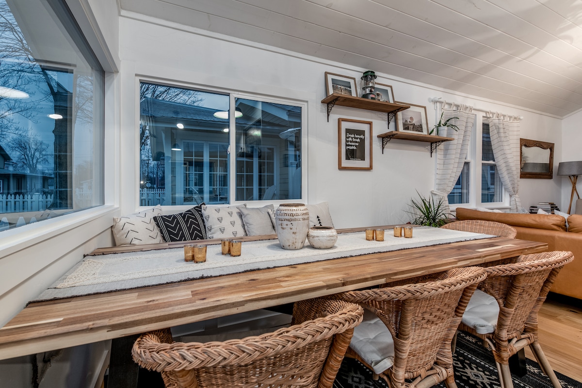 An open dining area features a long wooden table surrounded by woven chairs. Natural light streams through large windows, illuminating decorative touches such as potted plants and framed wall art. Soft curtains adorn the windows, enhancing the inviting feel of the space.