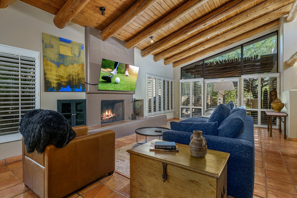 A spacious living area features a vaulted wood ceiling and terracotta tile flooring. A large flat-screen TV is mounted above a fireplace, while sliding glass doors open to outdoor views. Comfortable seating is arranged around a coffee table, creating a welcoming space.