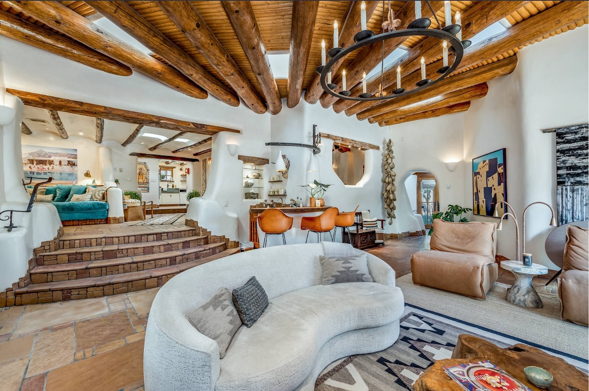 An open and airy living space showcases curved, plush seating and leather armchairs. Exposed wooden beams span the ceiling, with a rustic chandelier providing soft illumination. Textured rugs add warmth to the terracotta tile floor, inviting relaxation and conversation.