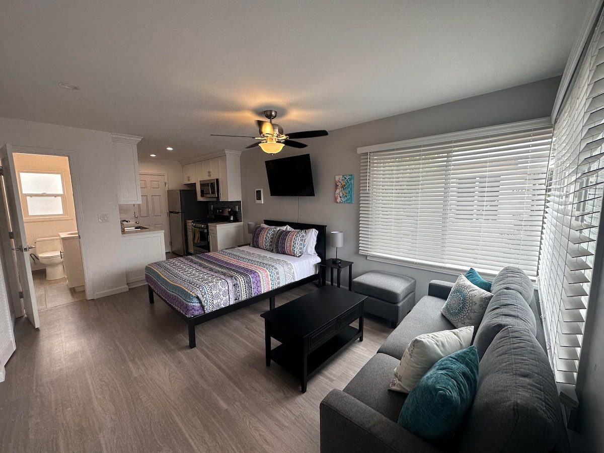 A well-lit studio apartment features a spacious bed adorned with colorful bedding. A comfortable gray sofa and a small coffee table are arranged in the living area. A modern kitchenette is visible in the background, along with a bathroom door that opens to a tiled space.