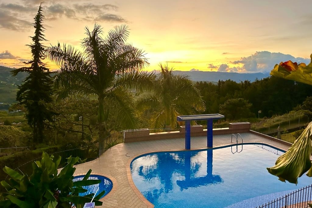 A tranquil swimming pool is surrounded by lush palm trees, reflecting the warm colors of the sunset. The pool area features a diving platform and is enclosed by a fence, offering views of the expansive natural landscape beyond.