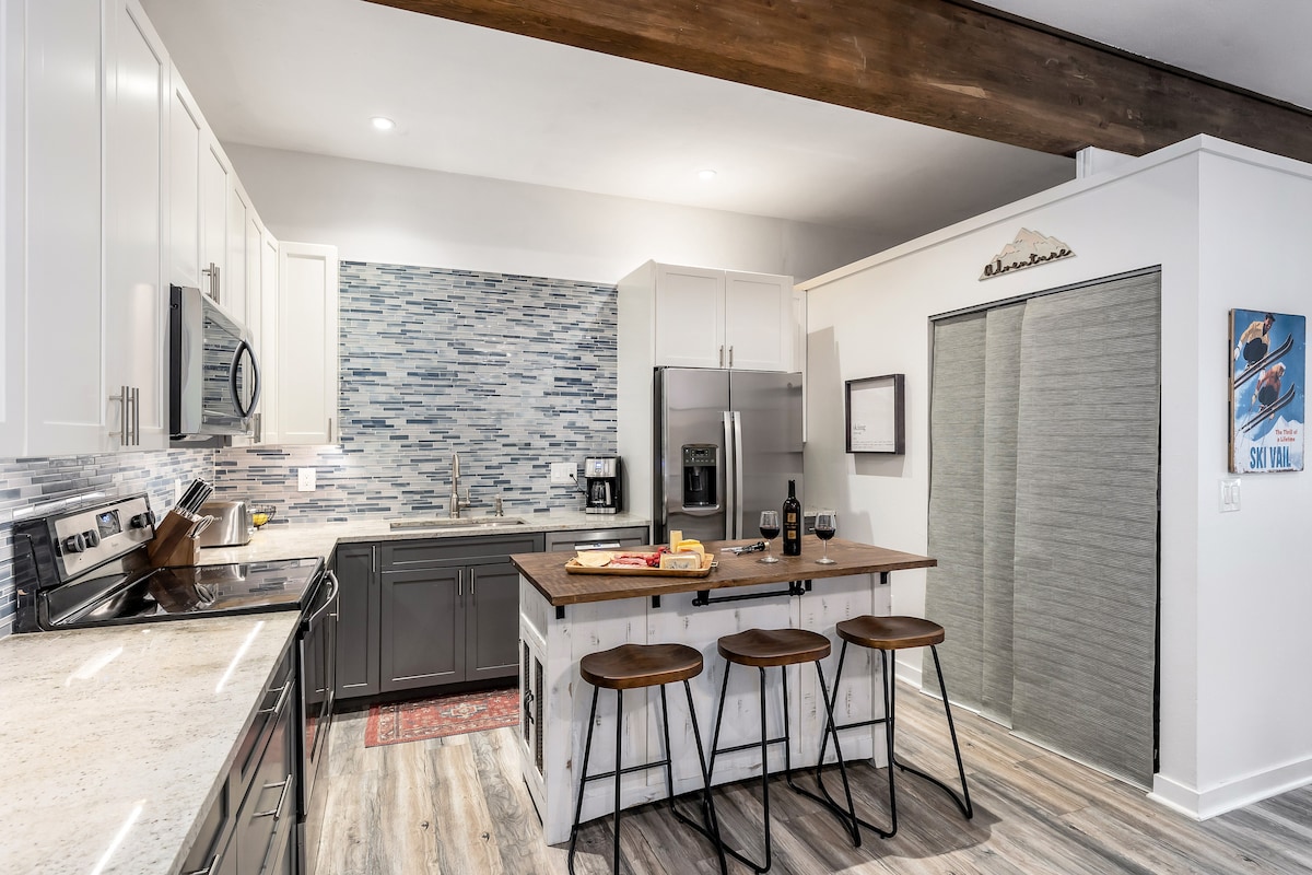 A modern kitchen features stainless-steel appliances, quartz countertops, and a central butcher block island with three bar stools. Light cabinets contrast with dark cabinetry, and a stylish backsplash adds visual interest to the space. Essential kitchenware and bottles are arranged on the countertop.