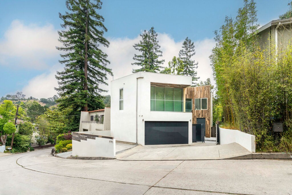 A modern two-story home is set against a backdrop of lush trees, showcasing a blend of wood and white exterior. The driveway leads to a garage, and the surrounding landscape features greenery, adding to the serene ambiance of the location.