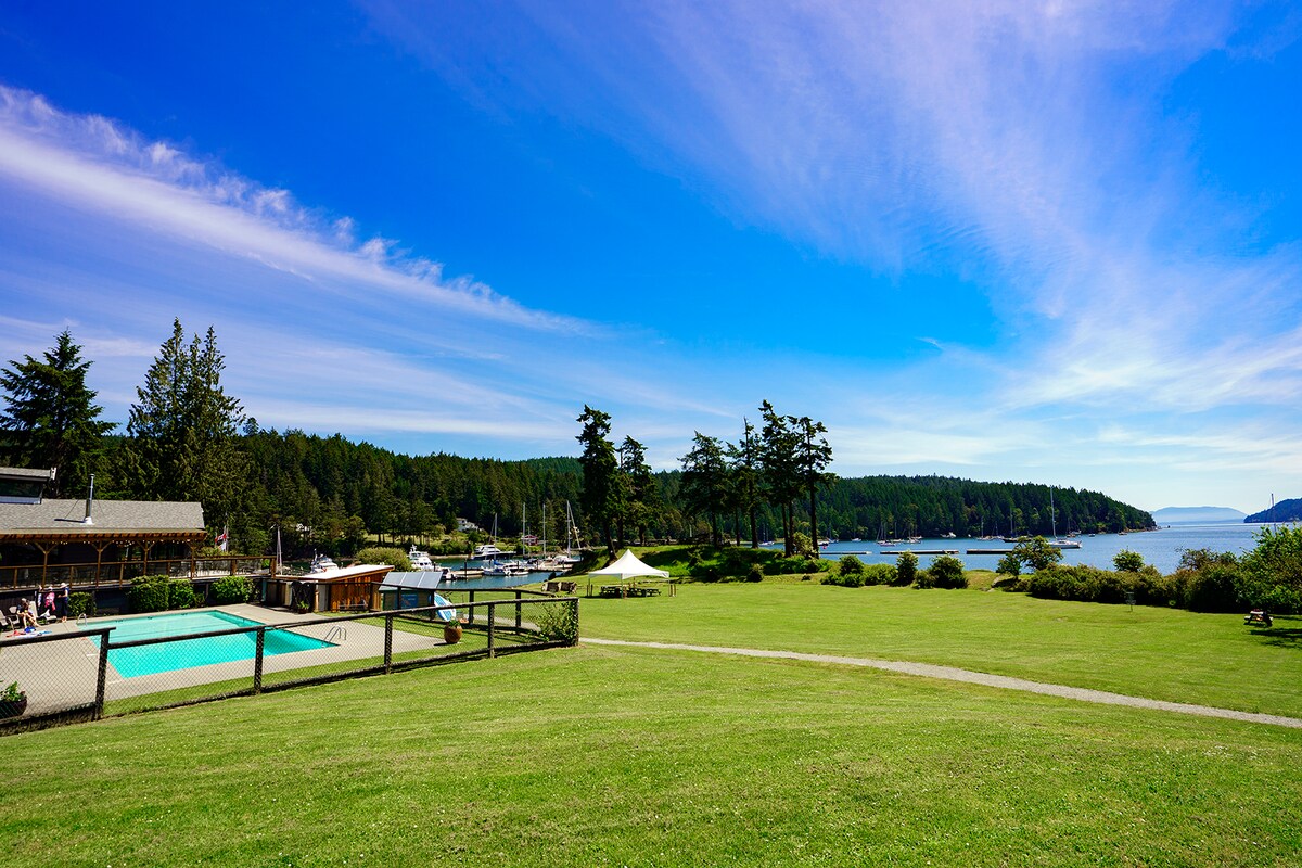 An expansive green lawn extends towards a tranquil body of water, flanked by trees and rolling hills. A swimming pool is visible near a building, with clear blue skies stretching overhead. The scene indicates a serene natural environment, perfect for relaxation and outdoor activities.