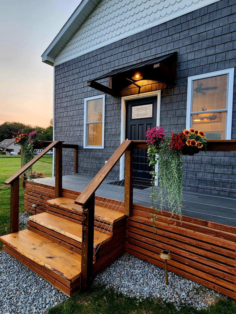 The entrance features a set of wooden steps leading up to a dark door, framed by two windows displaying decorative plants. Flowering pots hang from the railing, adding color to the exterior, with a warm light illuminating the entryway.