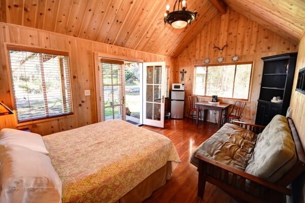 The cabin interior features warm wood paneling and spacious windows allowing natural light. A full-size bed is positioned beside a futon couch, with a dining table and kitchenette area visible. Air conditioning is present, and a small refrigerator stands nearby.