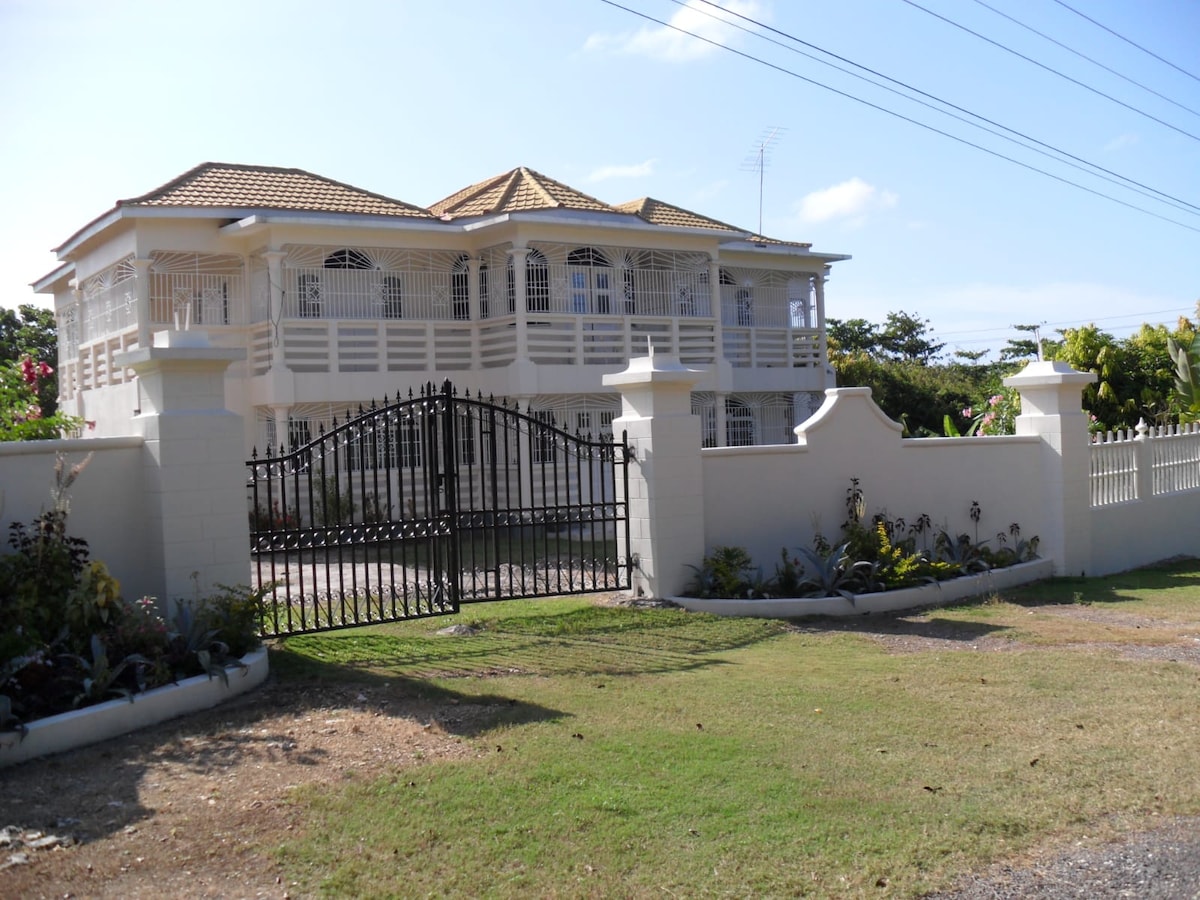 A two-story white villa is showcased, featuring a double-gabled roof and expansive balconies with decorative railings. A wrought iron gate stands at the entrance, framed by manicured lawns and surrounding greenery. Clear blue skies complete the serene residential setting.