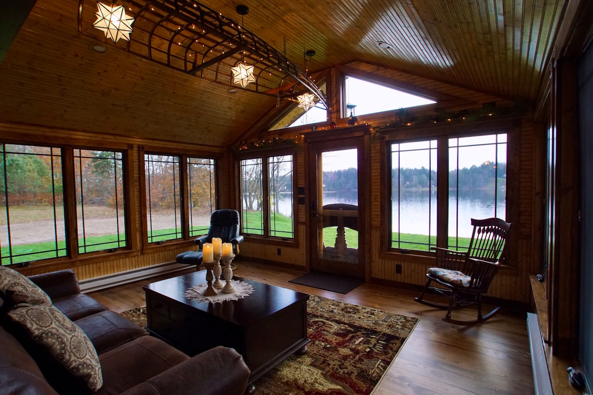 A sunlit sitting area features large windows that overlook the serene waterfront. A dark leather couch and a rocking chair offer seating options, while a coffee table with candles stands at the center. A warm wood ceiling adds to the inviting atmosphere.