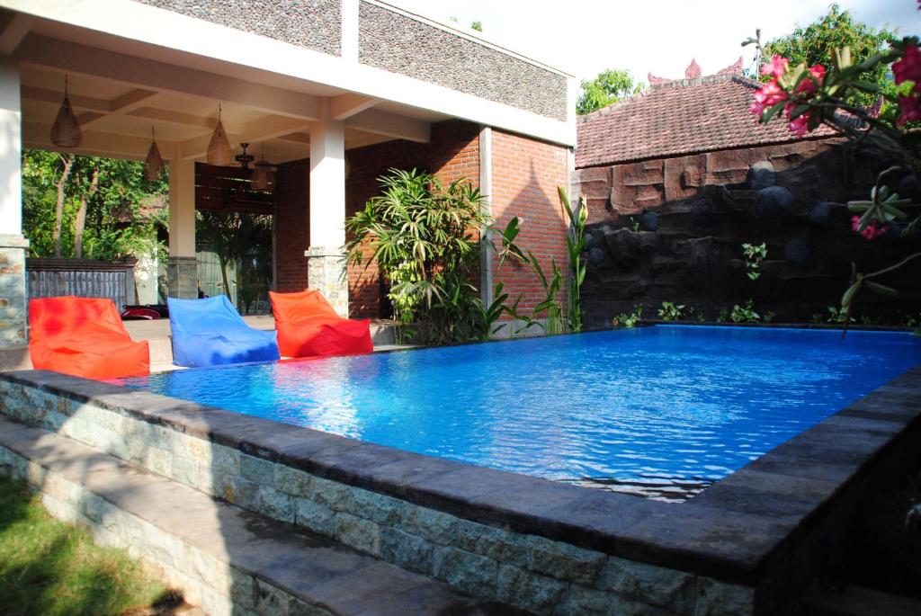 A serene pool area features clear blue water surrounded by lush greenery. Colorful lounge chairs in red and blue are positioned nearby, inviting relaxation. The space is well-integrated with the outdoor environment, showcasing natural stone accents and a charming structure in the background.