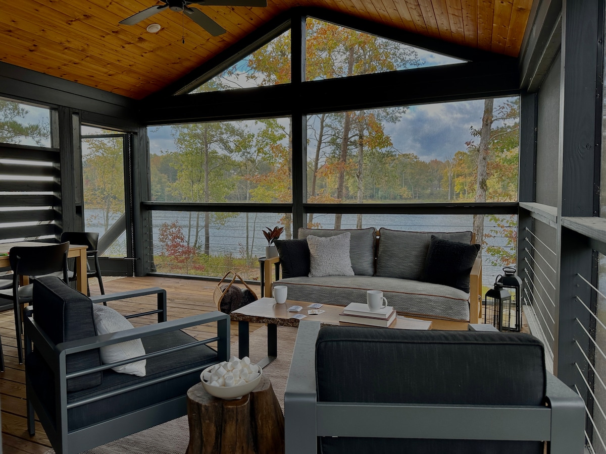 An inviting outdoor living space is shown, featuring comfortable seating arrangements on a spacious deck. Large windows frame serene lake views, while autumn foliage adds warmth. A rustic coffee table sits at the center, accompanied by a decorative lantern and a bowl of decorative items.