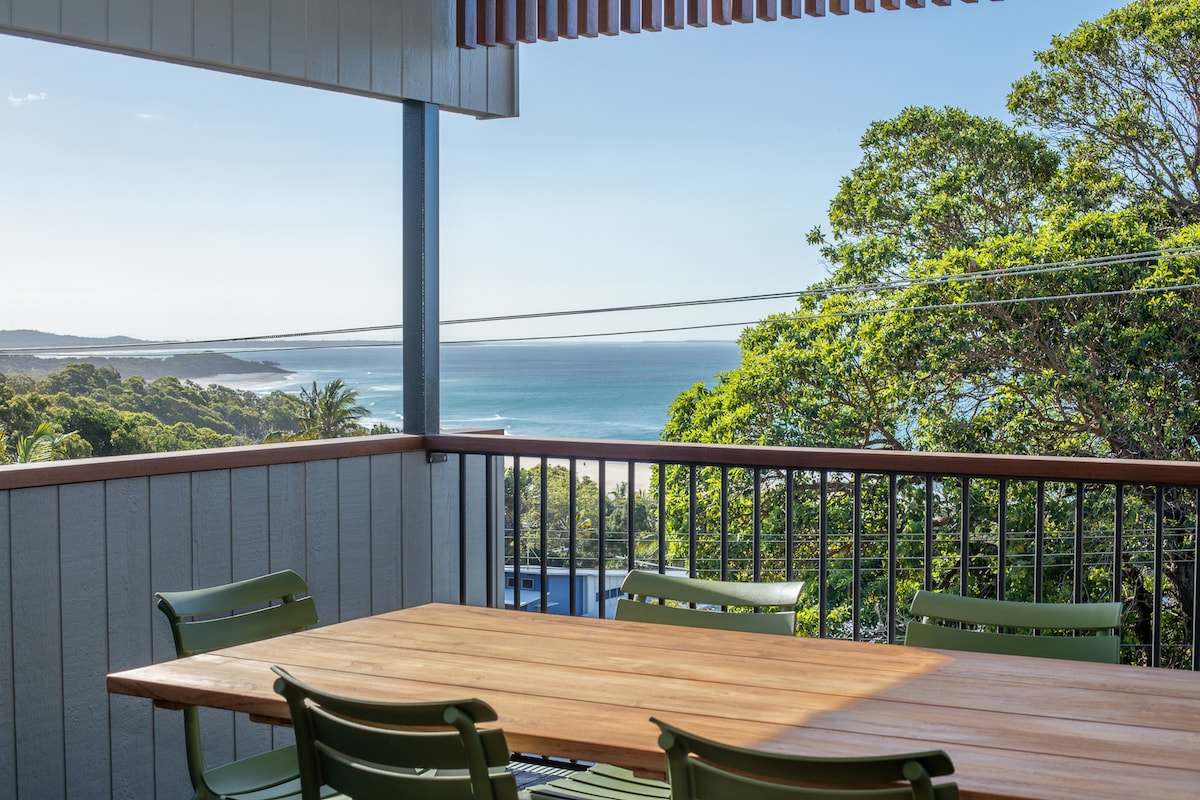 An outdoor dining area is presented with a large wooden table surrounded by six green chairs. Ocean views are visible in the distance through the lush surrounding trees, offering a serene backdrop to the space.