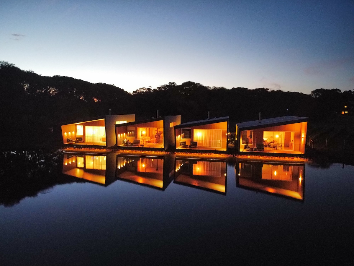 Three modern villas are illuminated at dusk, reflecting warmly on the tranquil lake's surface. Large glass windows reveal inviting indoor spaces, while the surrounding forest adds a serene backdrop to the retreat's enchanting evening ambiance.