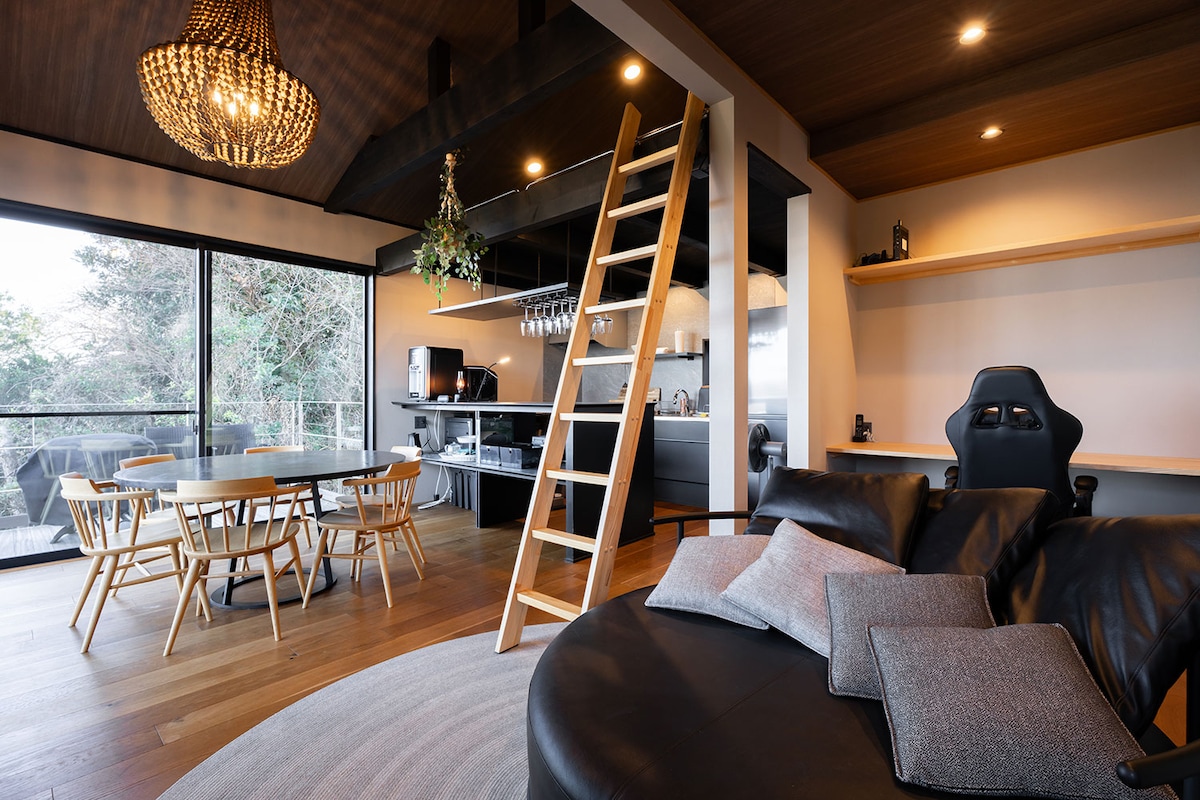 A modern living space is featured, showcasing a circular, plush sofa with neatly arranged cushions. A wooden ladder leads to a loft area, while a dining table is surrounded by six chairs. Large glass doors open to views of the surrounding greenery.