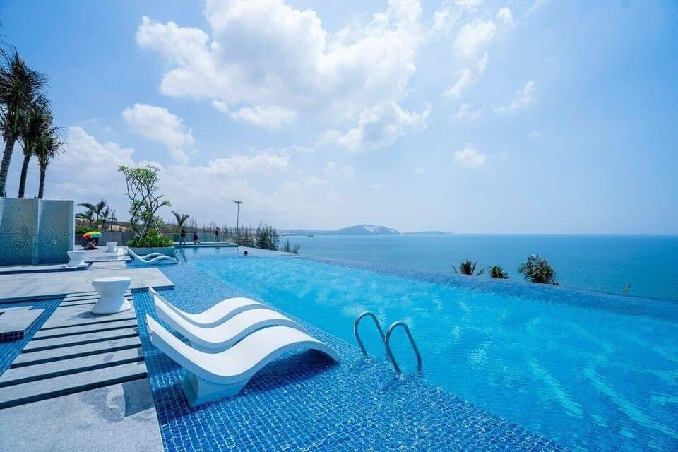 An inviting infinity pool stretches towards the horizon, offering stunning views of the ocean. Sun loungers beside the pool provide a space for relaxation, while palm trees sway gently in the breeze. The expansive blue sky enhances the serene atmosphere of the location.