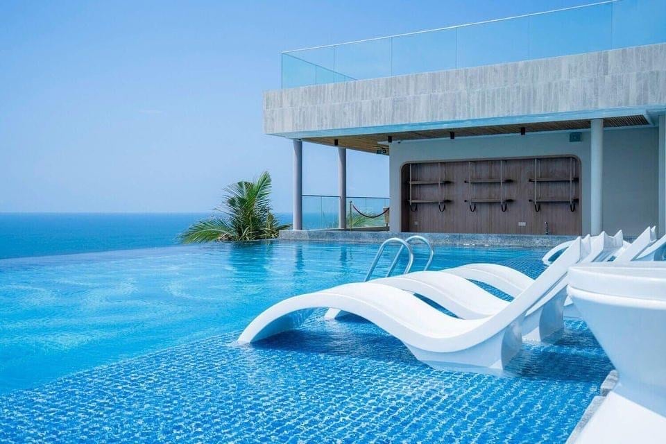 A stunning infinity pool is situated on a rooftop terrace, overlooking a vast ocean. Sun loungers are positioned along the pool's edge, inviting relaxation. The clear blue sky complements the serene waters, enhancing the tranquil ambiance of the space.