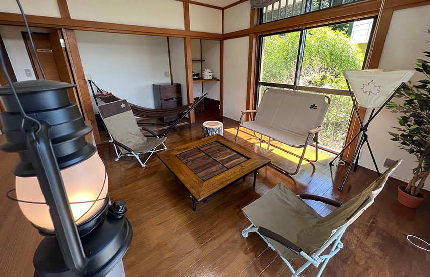 An indoor camping room features two comfortable folding chairs and a lounge chair arranged around a wooden coffee table. A lantern is situated in the foreground, and a hammock is seen hanging nearby. Large windows offer views of the greenery outside.