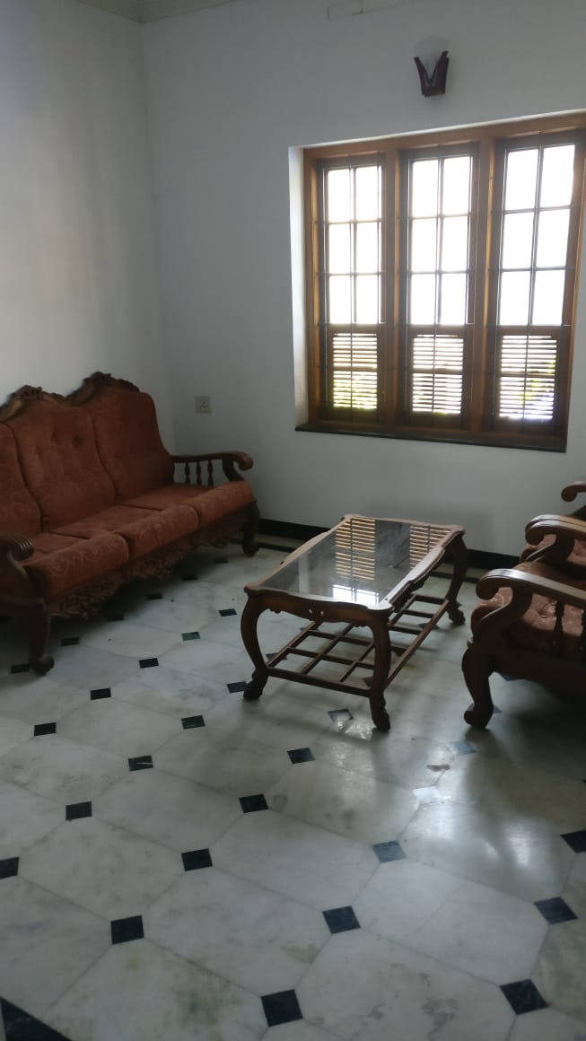 A seating area features a pair of traditional armchairs and a matching sofa, all upholstered in a rich, textured fabric. A glass-topped coffee table rests on a patterned marble floor, while a window with wooden shutters allows natural light to fill the space.