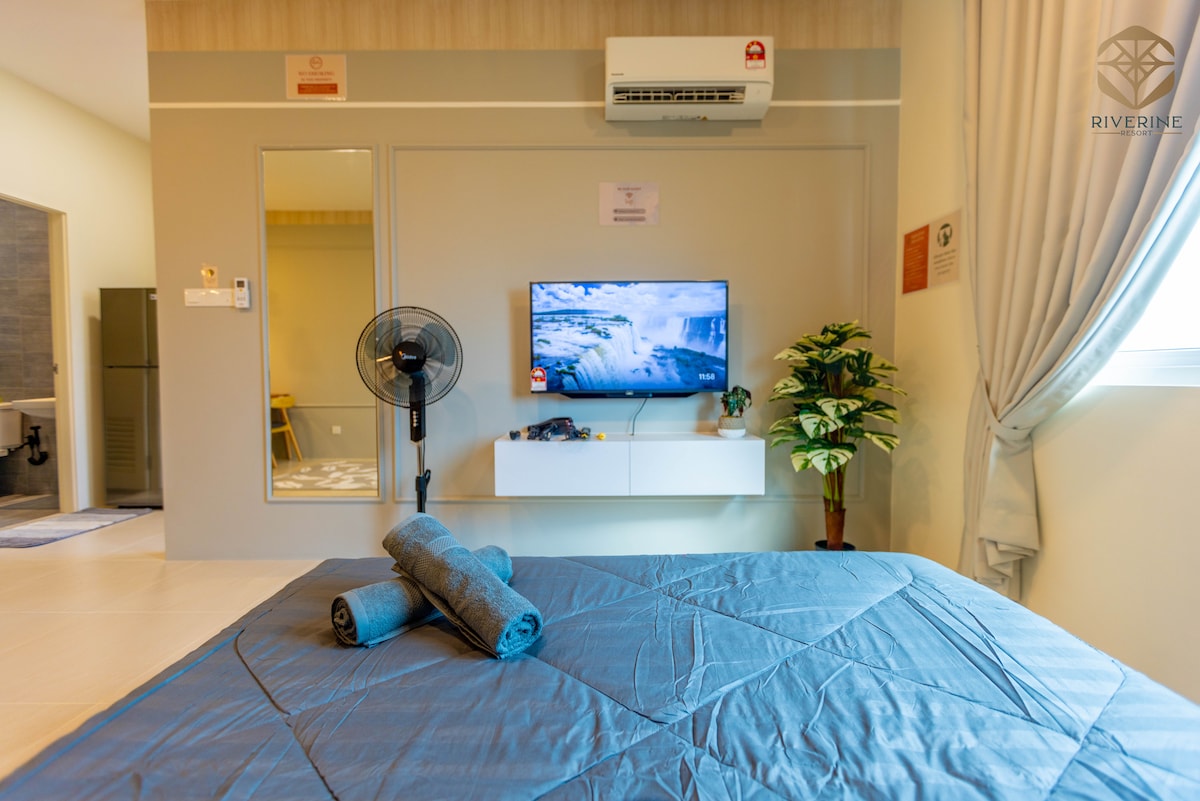 A studio room is shown with a comfortable bed covered in a blue quilt. A television is mounted on the wall opposite the bed, with a minimalistic white cabinet below it. A fan and a potted plant are positioned nearby, enhancing the room's functionality.
