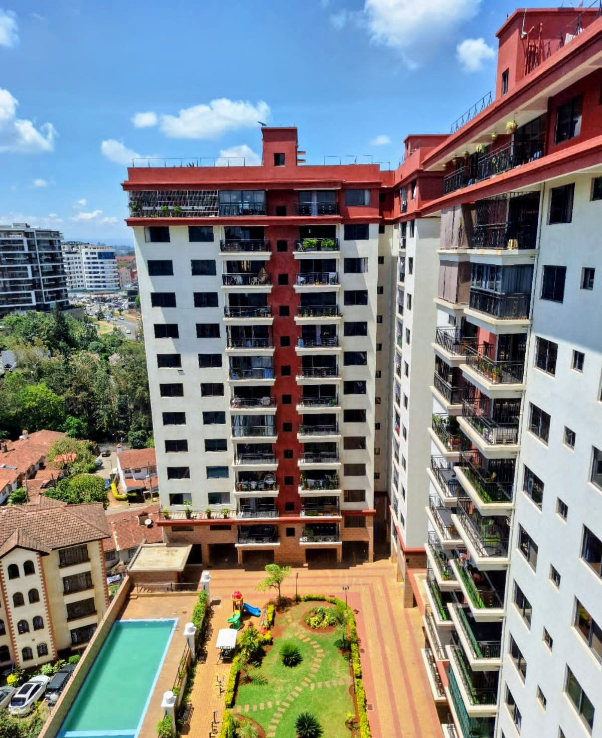 A multi-story building is showcased from a high vantage point, revealing multiple balconies adorned with greenery. A swimming pool is visible in the foreground, surrounded by colorful landscaping, with leisure areas designed for relaxation and play.