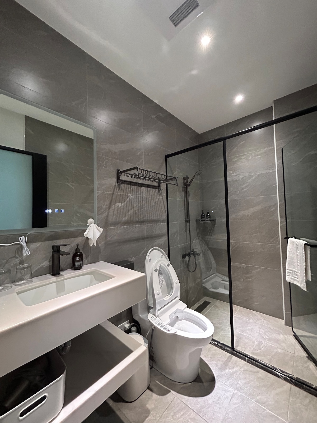 A modern bathroom features sleek gray tiles and a spacious walk-in shower with a glass enclosure. A white vanity with an integrated sink is accompanied by a large mirror, while essentials are neatly arranged. Towels and toiletries are available for convenience.