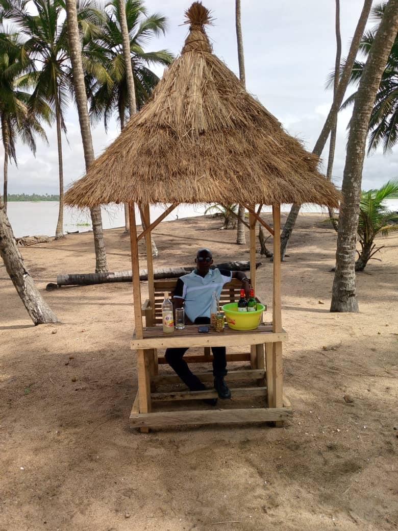 A shaded wooden gazebo stands on sandy ground, surrounded by palm trees. A person is seated at a rustic table with a bowl of fresh fruits and drinks. The tranquil water can be seen in the background, enhancing the serene environment.