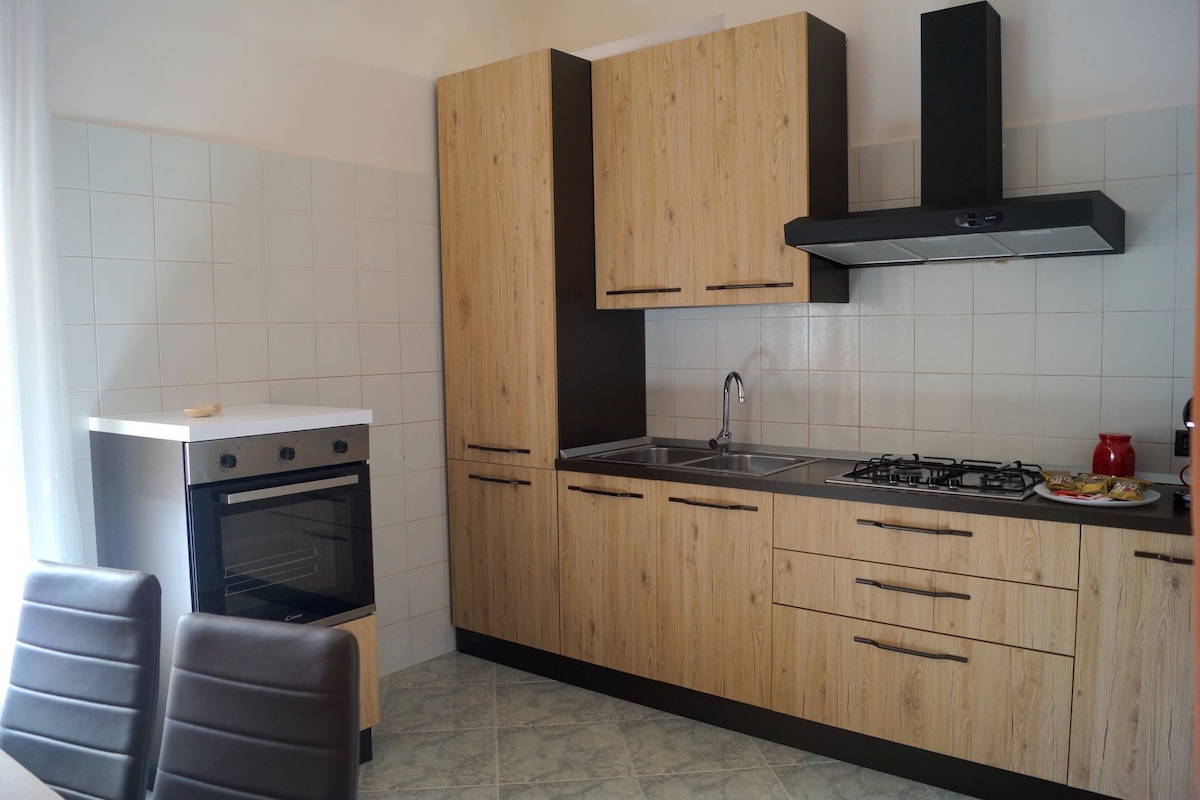A modern kitchen features wood-grain cabinetry and sleek black accents. Stainless steel appliances include an oven and a gas stove. A kitchen sink is positioned between the countertops, complemented by a window allowing natural light to brighten the space. A dining table with chairs is visible.