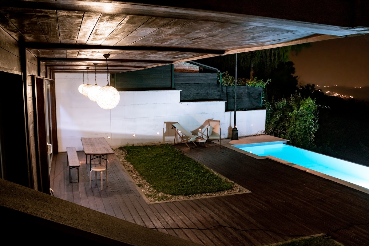 An outdoor area is featured, featuring a wooden deck with a long dining table and chairs. Soft lighting illuminates the space, while a pool with a cool blue hue is positioned nearby. A grassy area and a lounge chair enhance the inviting setting.