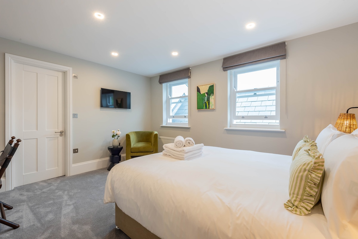 A spacious bedroom is equipped with a luxurious king bed layered with fresh linens. A cozy armchair sits in the corner, while a smart TV is mounted on the wall. Natural light enters through two windows, alongside a simple wardrobe and decorative wall art.