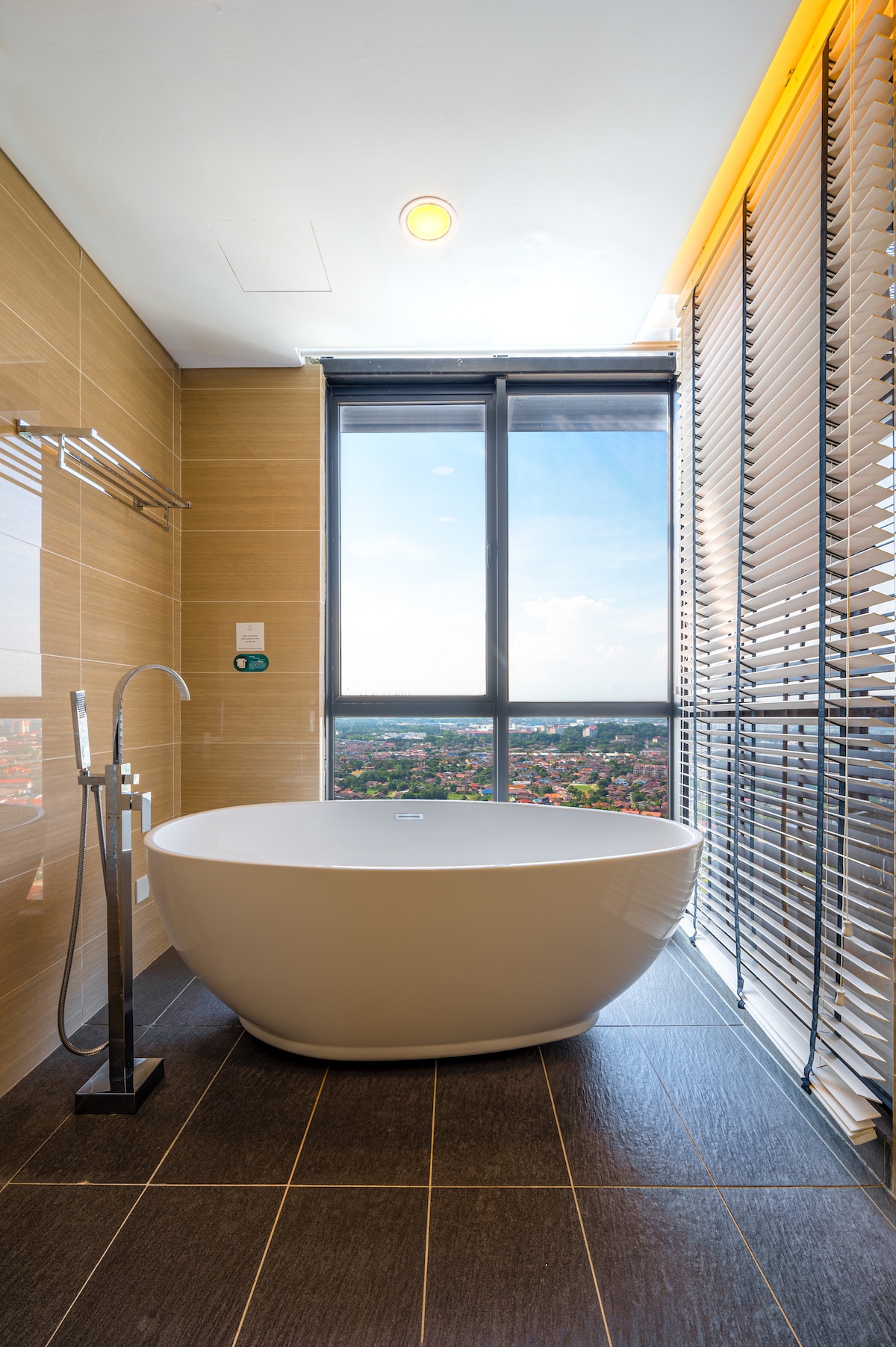A modern bathroom features a spacious freestanding bathtub positioned near a large window, offering an expansive view of the surroundings. Natural light floods the space through the glass, complemented by stylish blinds. Neutral-toned tiles and sleek fixtures contribute to a contemporary ambiance.