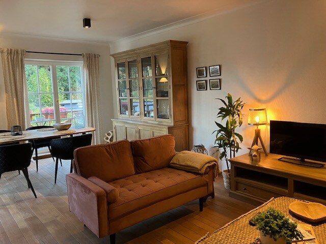 A spacious living area features a comfortable sofa with a throw blanket, complemented by a wooden side table. A dining space is visible with a table and chairs. Large glass doors lead to a terrace, allowing natural light to fill the room.
