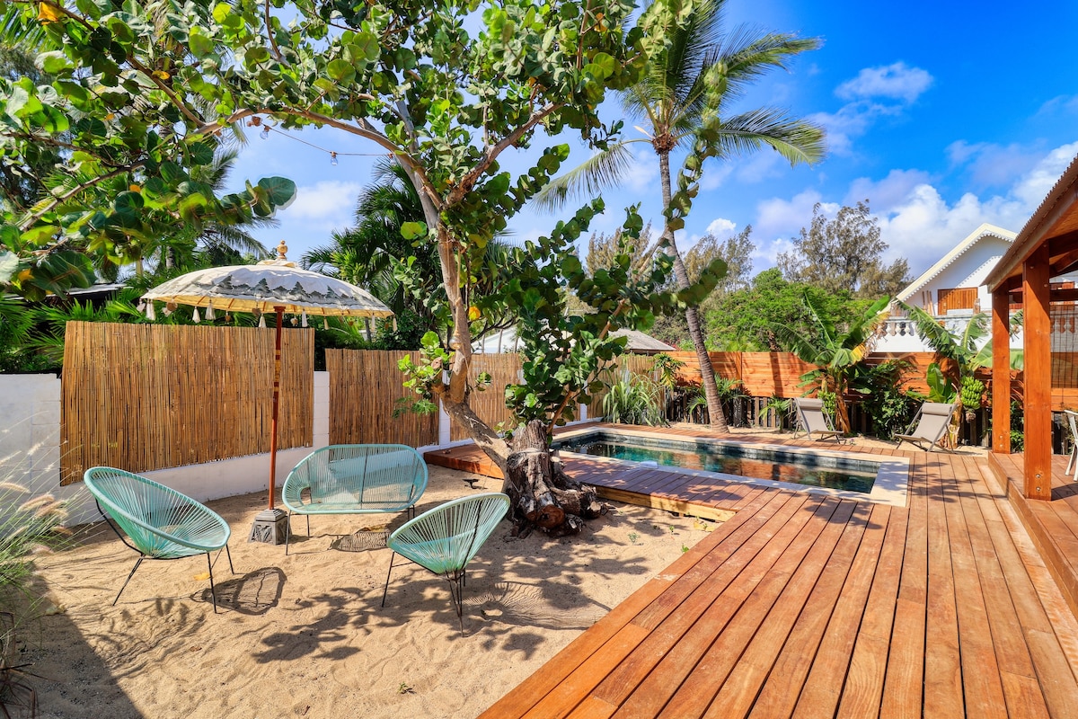 A serene outdoor space features a private pool surrounded by a wooden deck. Comfortable seating is arranged on sandy ground under the shade of trees. A thatched umbrella enhances the relaxation area, while tall plants provide privacy from neighboring properties.