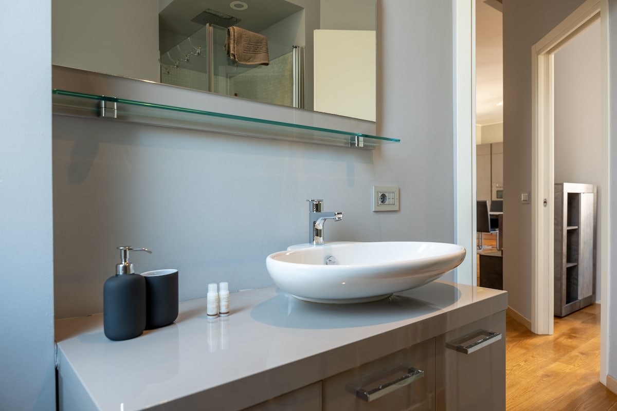 The bathroom features a stylish countertop with a circular sink, complemented by a modern faucet. A clear glass shelf above holds personal items. The space is illuminated by natural light, highlighting the sleek design of the cabinetry and the warm wooden flooring.