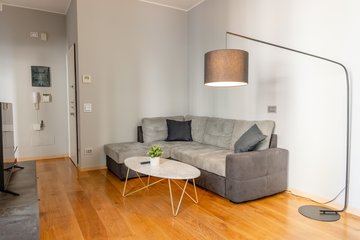 A spacious living area is presented with a large sectional sofa in soft gray, complemented by a chic floor lamp. A light wood table sits at the center, accompanied by a small green plant for a touch of color. Natural light enhances the warm wooden flooring.