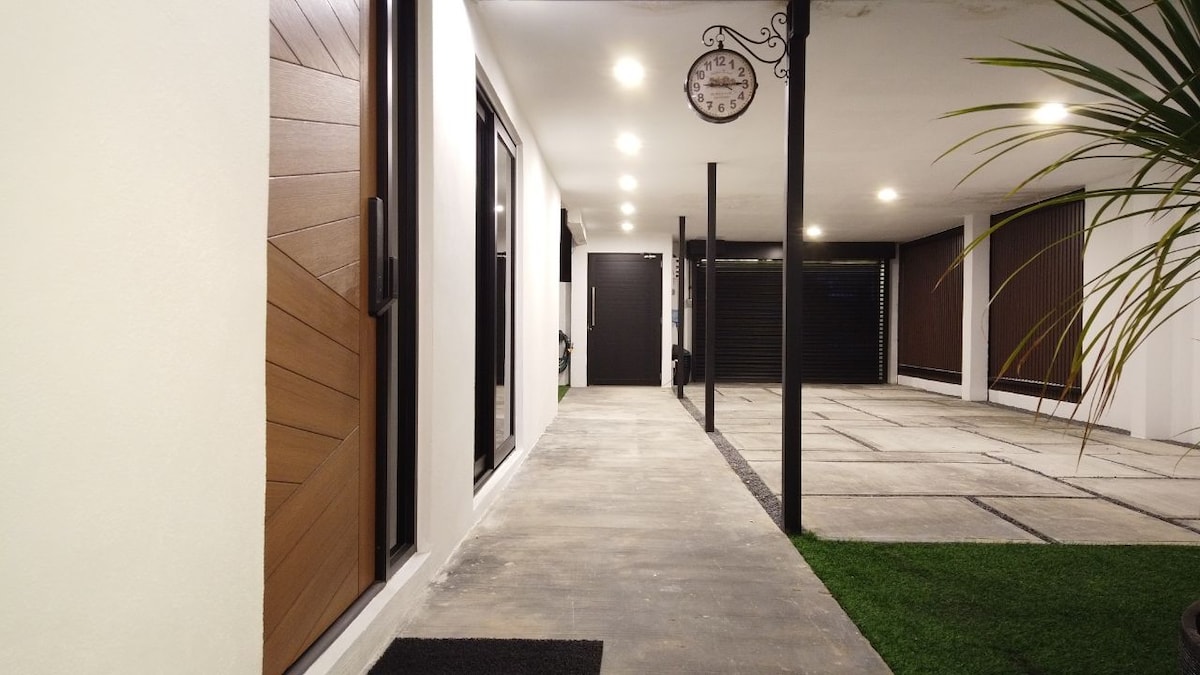 A well-lit passageway is flanked by smooth white walls and wooden doors, leading to garage parking on the right. The modern clock adds a touch of charm, while the landscaped area features greenery alongside the stone pathway.