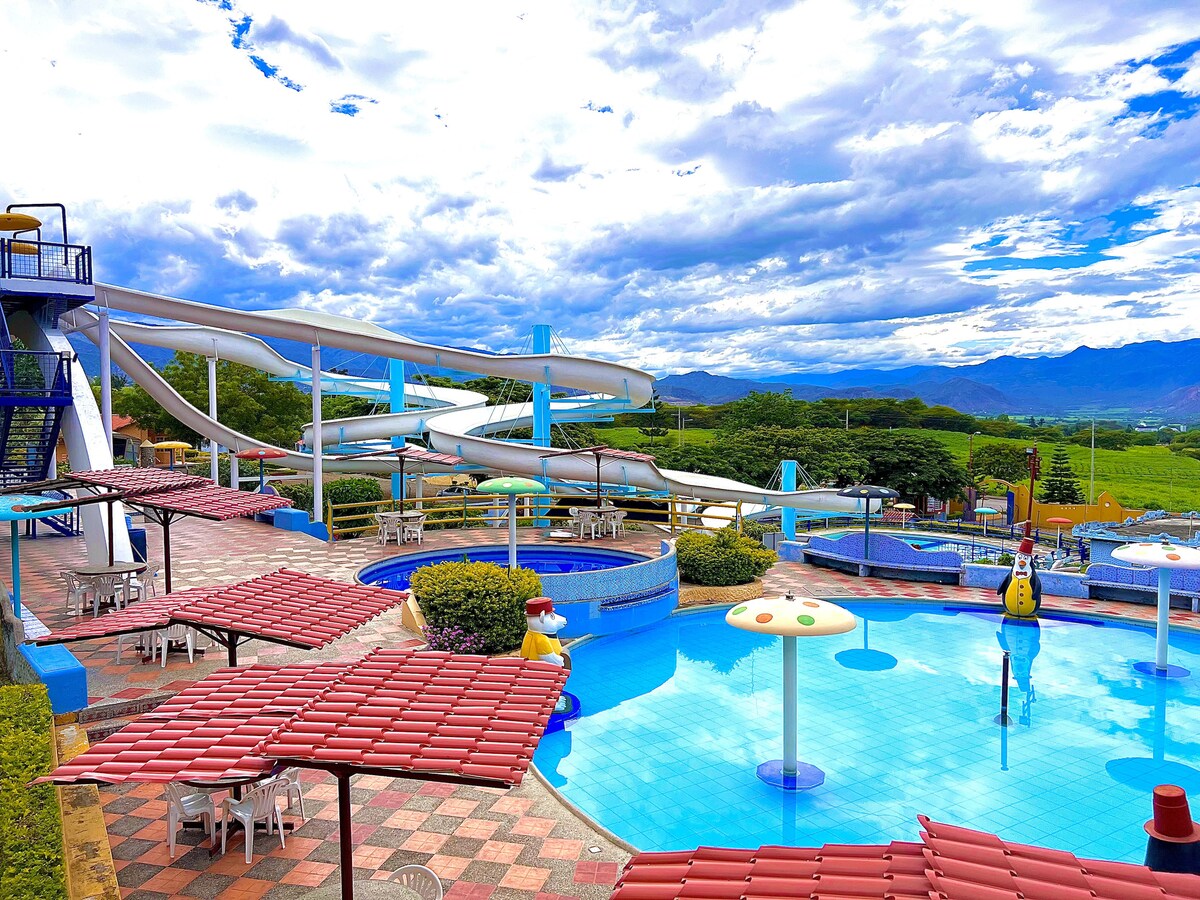 A water park area is depicted, featuring multiple water slides and a swimming pool. Sun loungers and tables with umbrellas surround the pool, offering shaded seating. Lush greenery and mountains can be seen in the background under a cloudy sky.