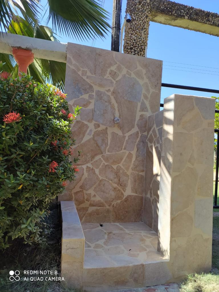 An outdoor shower stall is crafted from light-colored stone, featuring a natural rock pattern. It is set against a backdrop of greenery, with vibrant flowers visible nearby. The shower includes a horizontal bench and a simple overhead fixture.