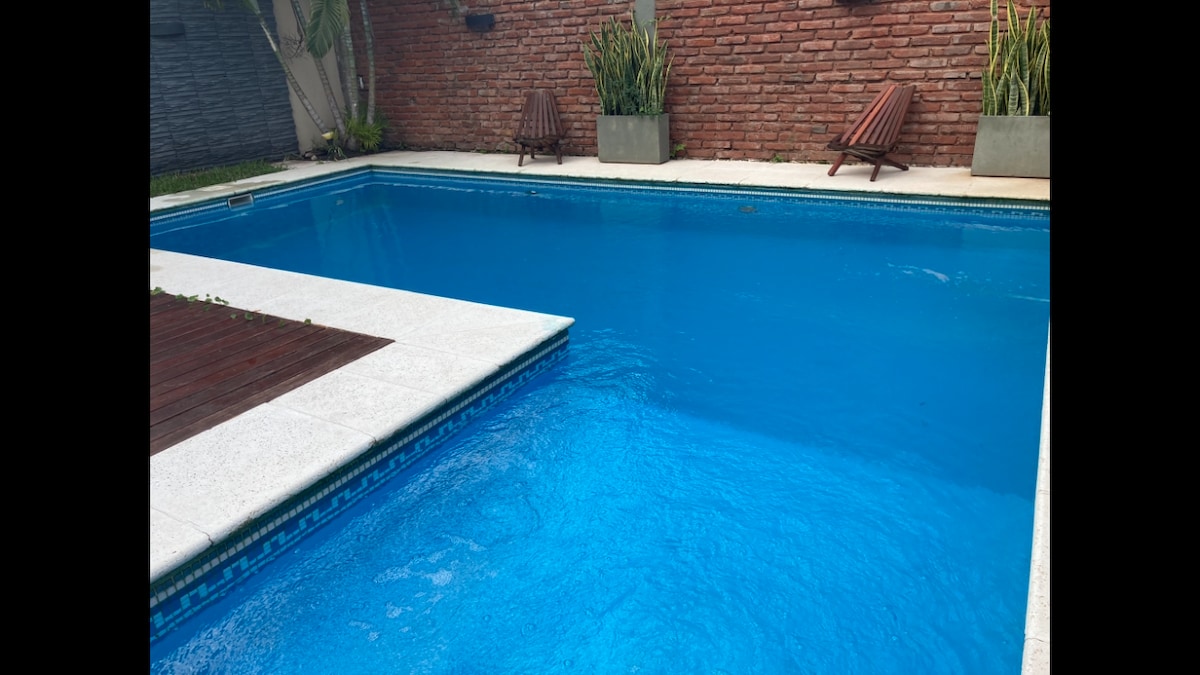 A tranquil swimming pool is surrounded by a paved deck, featuring light-colored tiles. Wooden lounge chairs are placed on one side, while tall planters with greenery add a touch of nature. The water is a clear blue, inviting relaxation.