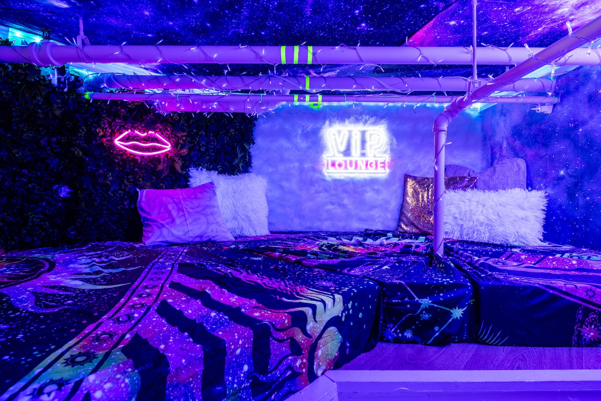 A cozy upper-level lounge area features vibrant galactic decor, including a deep blue ceiling and neon 'VIP Lounge' sign. Plush pillows and a richly patterned blanket create a comfortable space, surrounded by fuzzy walls and ambient lighting with black lights to enhance the playful atmosphere.