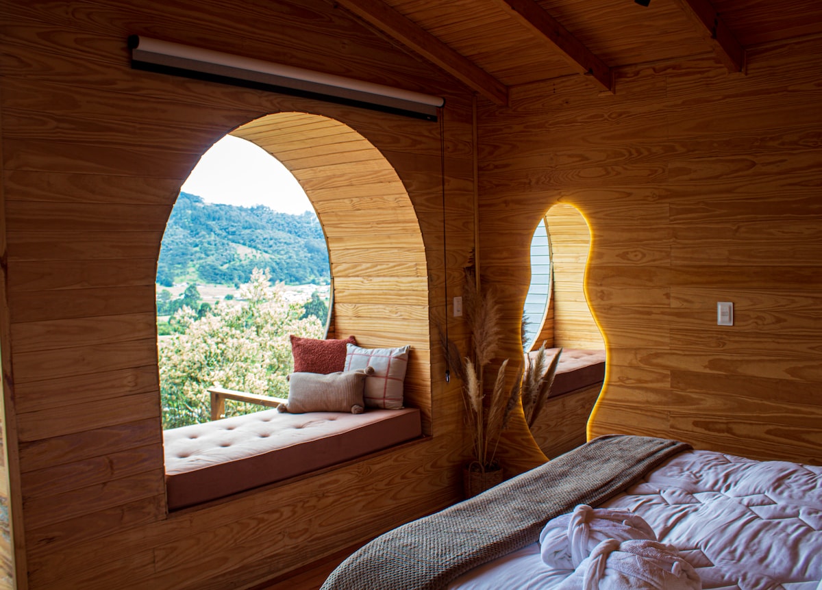 A spacious sleeping area is adorned with wooden walls, featuring a cozy king-size bed. Unique arched windows provide views of the surrounding landscape, with a cushioned bench beneath one window and soft lighting by the other, enhancing the tranquil ambiance.
