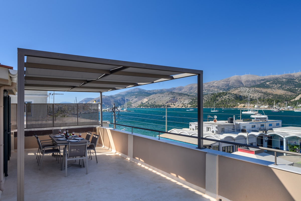 A spacious terrace features a large dining table surrounded by chairs, with sun loungers positioned nearby. The area is shaded by a pergola, offering sweeping panoramic views of the sea, nearby hills, and boats in the harbor, creating an inviting outdoor space.