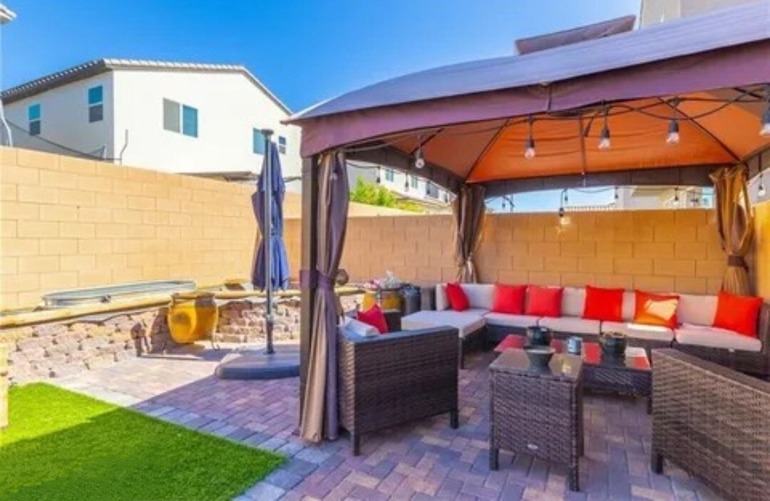 An outdoor seating area is displayed, featuring a covered gazebo with a series of string lights. Comfortable wicker furniture is arranged around a low table, accented with colorful cushions. Artificial grass and stone pavers create a welcoming space for relaxation.