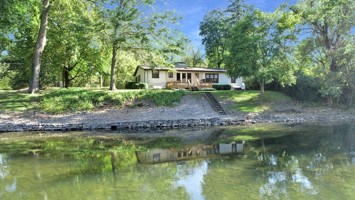 A single-story house is situated along the Juniata River, surrounded by greenery. A wide deck extends from the structure, providing access to the outdoors. Stairs lead down to the water's edge, reflecting the home and trees in the calm river surface.