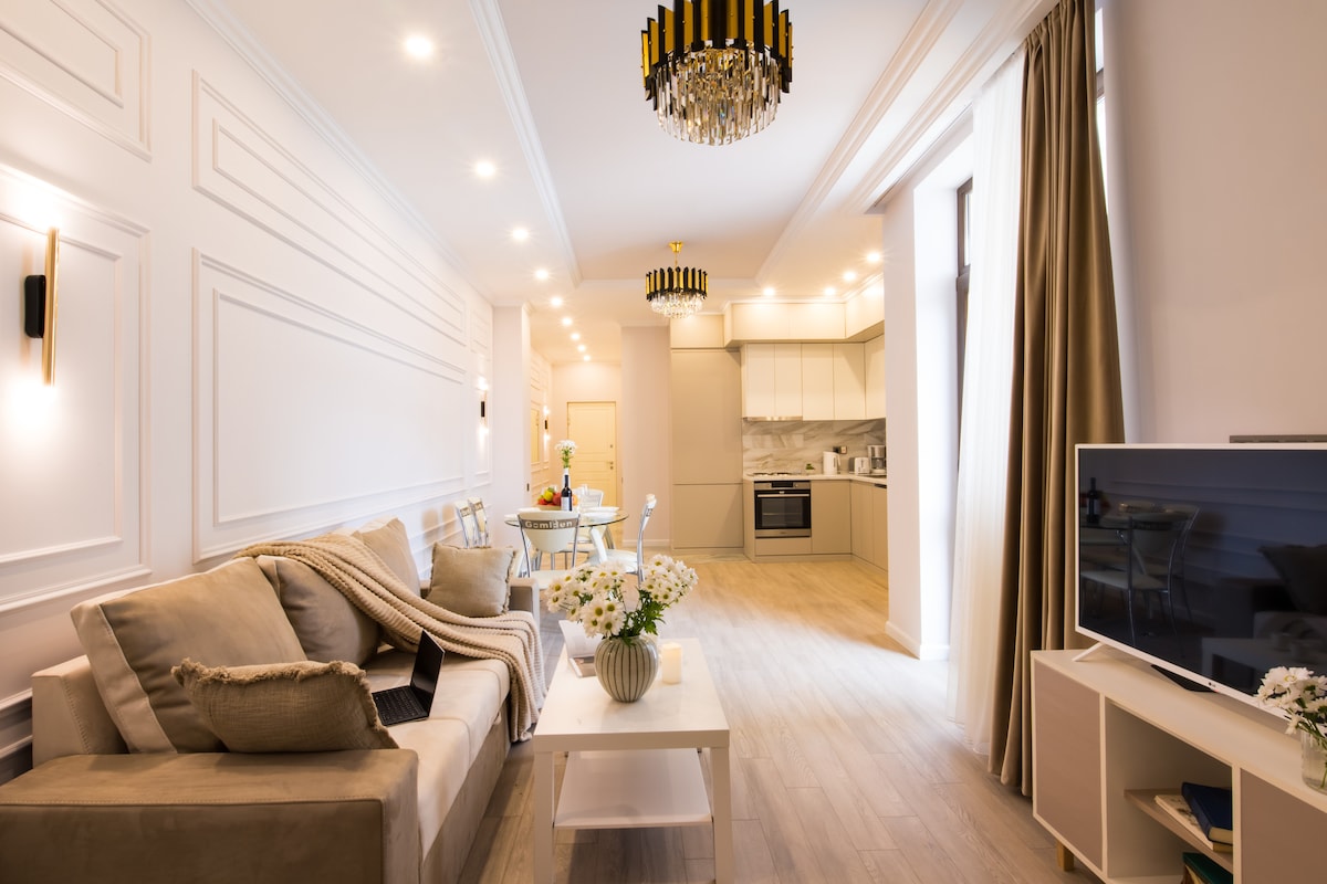 An open-plan living area features elegant decor with light-coloured walls and wooden flooring. A comfortable sofa is dressed with soft cushions and a throw blanket. A dining area is visible in the background, along with a modern kitchen. Bright lighting enhances the inviting atmosphere.