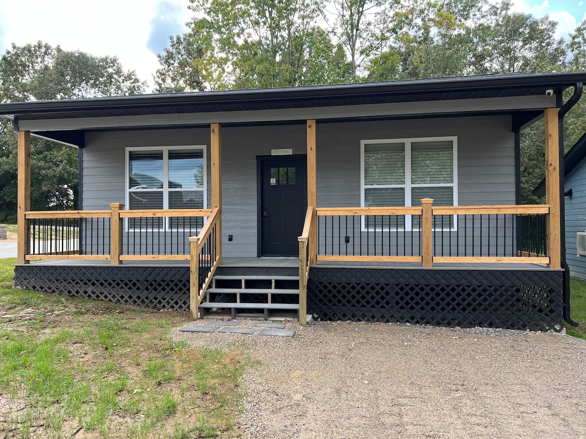 The exterior of a cabin features a welcoming front porch with wooden railings and steps leading to the entrance. Large windows allow natural light to fill the space, and the building is surrounded by a gravel area, framed by trees in the background.